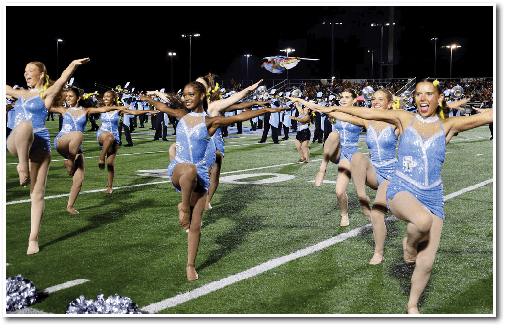 Adults in blue sparkly dance costumes perform a synchronized high-kick routine on a football field at night with a crowd and marching band in the background. Description generated by AI