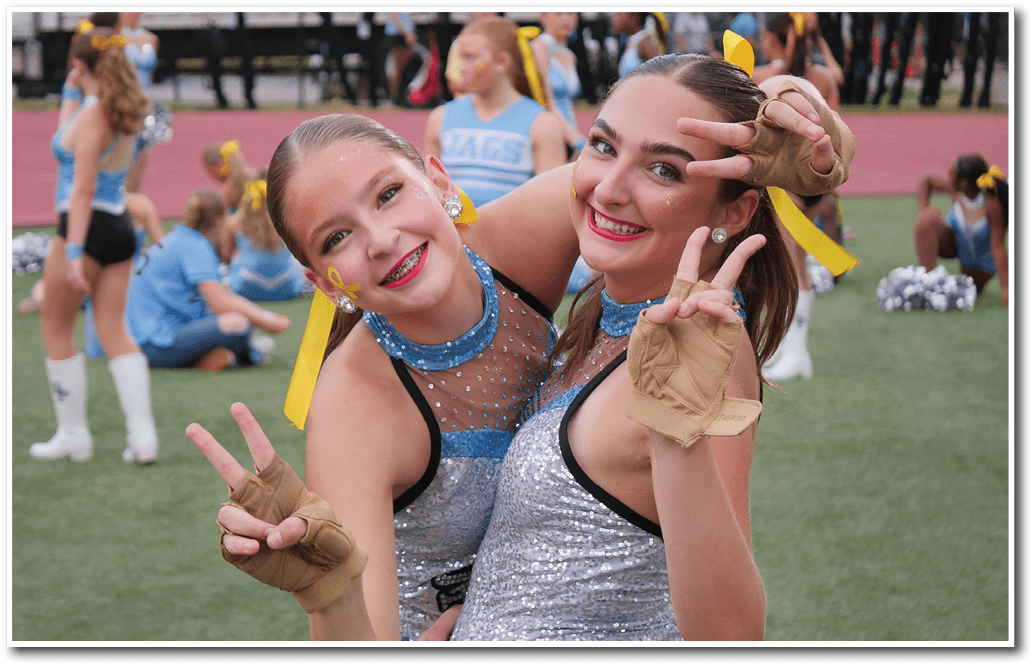 Two adults in sparkly cheerleading outfits with yellow ribbons and gloves smile and pose with peace signs on a sports field with other cheerleaders in the background. Description generated by AI