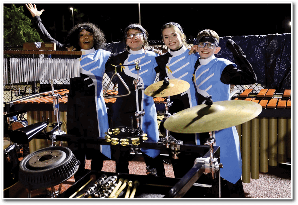 Four people in matching blue and black uniforms stand together smiling behind a percussion instrument setup at night. Description generated by AI