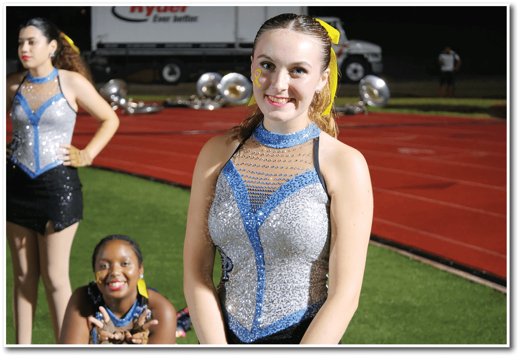 Three people wearing sparkly blue and silver performance outfits are on a field near a running track, with one person smiling at the camera and others posing behind. Description generated by AI
