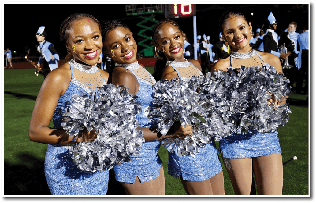 Four adults wearing sparkly blue dresses and holding silver pom-poms smile while standing on a grassy field at night with a marching band in the background. Description generated by AI