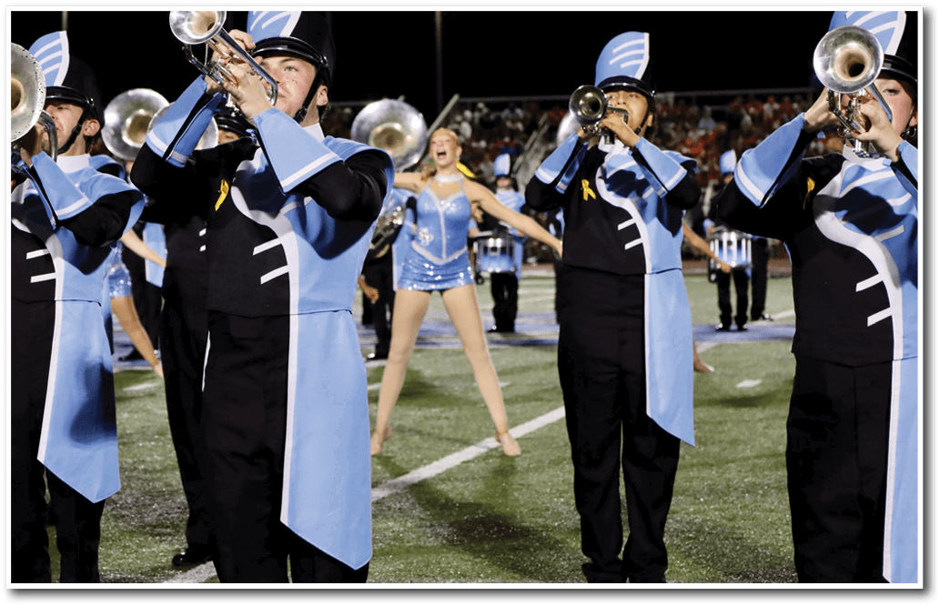 People in marching band uniforms stand on a field with a person in a blue sequined outfit visible in the background. Description generated by AI