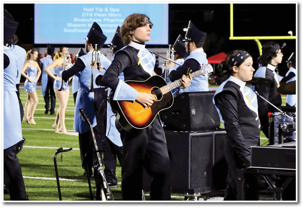 People in marching band uniforms are performing on a football field with one person playing an acoustic guitar and a scoreboard in the background showing a game between Spain Park and another team. Description generated by AI