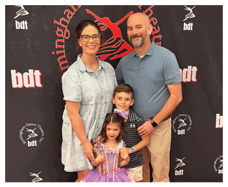 A group of four people, including two adults and two children, pose smiling in front of a backdrop with dance theater logos, with one child wearing a purple ballet costume. Description generated by AI