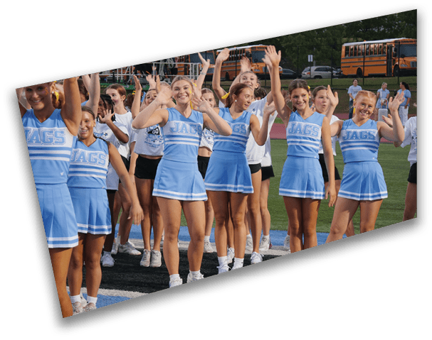A group of people in blue cheerleading uniforms with “JAGS" on the front are smiling and raising their hands outdoors on a field with school buses in the background. Description generated by AI