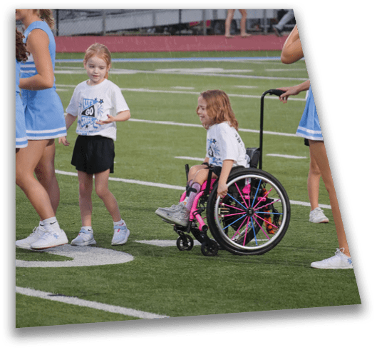 Children in sports attire are gathered on a field with one child in a wheelchair being pushed by another person. Description generated by AI