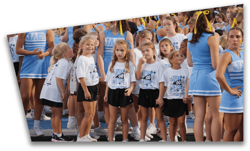 A group of children wearing matching cheerleading uniforms and t-shirts are gathered outdoors, some smiling and others looking around, with adults in blue cheerleader outfits nearby. Description generated by AI