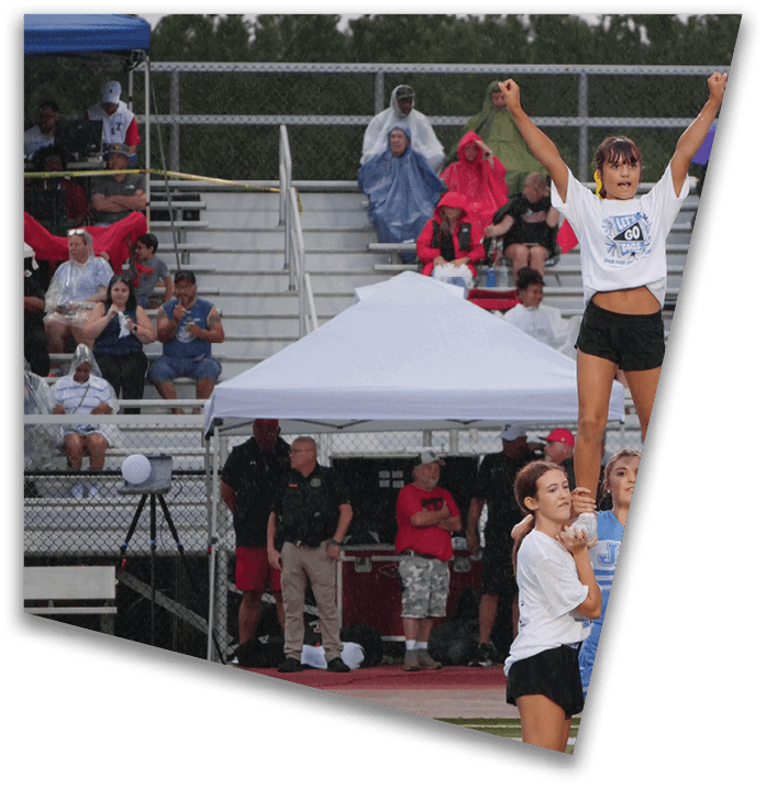 A group of people in casual clothing and rain ponchos watch from bleachers while a person in a white shirt and black shorts is lifted by others during an outdoor event. Description generated by AI