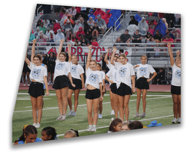 A group of children wearing matching white t-shirts and black shorts are performing a cheer routine on a sports field with a crowd watching from the bleachers. Description generated by AI