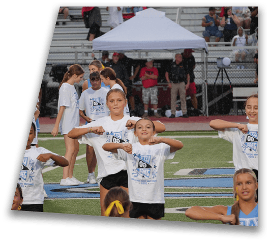 Children wearing matching white t-shirts are performing a coordinated dance routine on a sports field with bleachers and a tent in the background. Description generated by AI