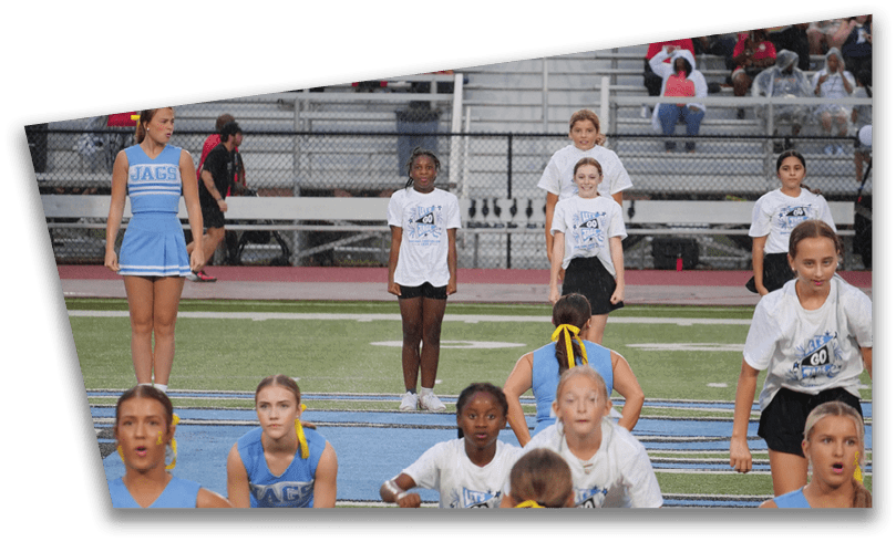 Children in cheerleading uniforms and sportswear are standing and sitting on a sports field with bleachers and spectators in the background. Description generated by AI