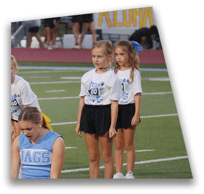 Children wearing cheerleading outfits stand on a sports field with one child in a blue uniform looking down and others in white shirts and black skirts standing behind. Description generated by AI