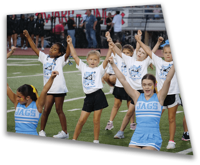 Children in sports uniforms are performing a cheer routine on a field with their arms raised. Description generated by AI