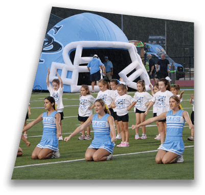 Children in sports uniforms and cheerleading outfits are performing a group routine on a field near an inflatable tunnel and a track. Description generated by AI