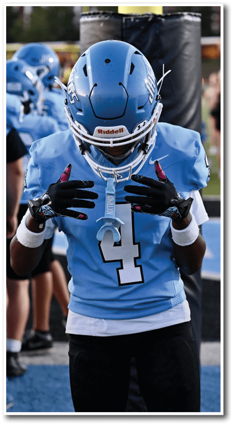 A person wearing a blue football uniform with the number 4 is adjusting their gloves on a field while other players in similar uniforms stand in the background. Description generated by AI