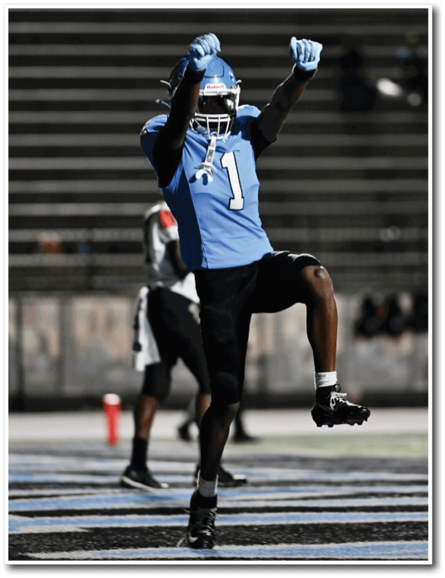 A person in a blue football uniform with the number 1 is celebrating with raised arms while other players and a football are visible on the field during a nighttime game. Description generated by AI