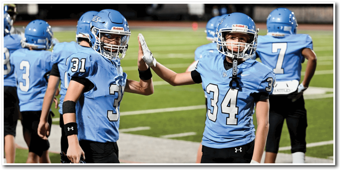 Two people wearing light blue football jerseys with numbers 31 and 34 are giving each other a fist bump on a football field. Description generated by AI