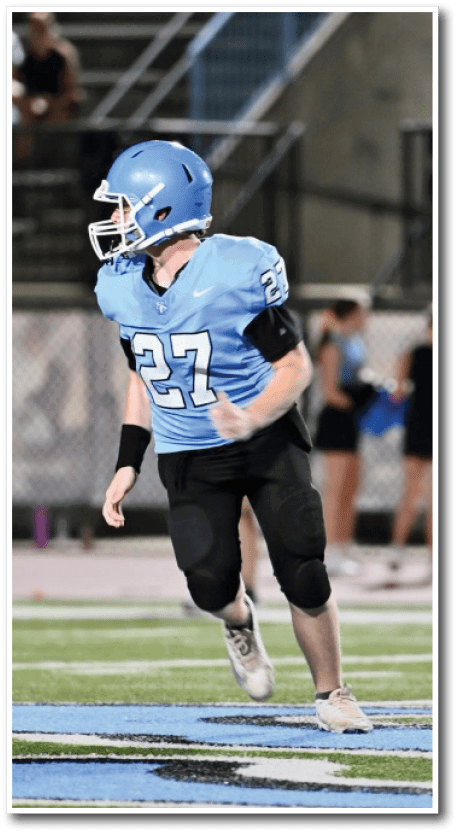 A person wearing a blue football uniform with the number 27 is running on a football field near the center logo with spectators in the background. Description generated by AI