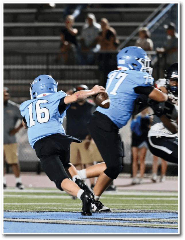 Two people in blue football uniforms are engaged in a play on a football field with one holding the ball and the other running, while a person in a black and white uniform approaches them. Description generated by AI