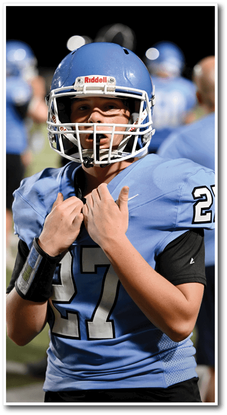 A person wearing a blue football uniform and helmet stands on a field with other similarly dressed people blurred in the background. Description generated by AI