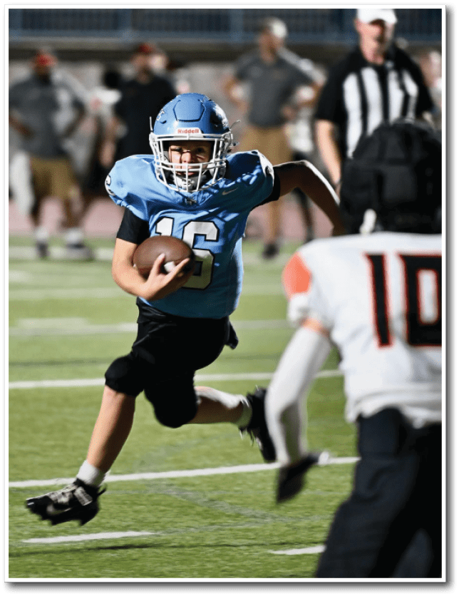 A person in a blue football uniform is running with the ball on a field while two people in white and black uniforms approach to block or tackle. Description generated by AI