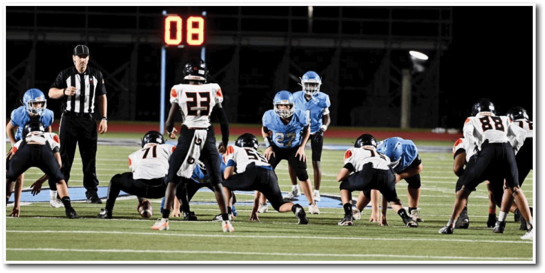People in football uniforms are lined up on a field facing each other, preparing for a play during a game. Description generated by AI