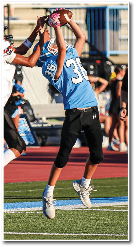 Two people in football uniforms are jumping to catch a football on a field with a referee and spectators in the background. Description generated by AI