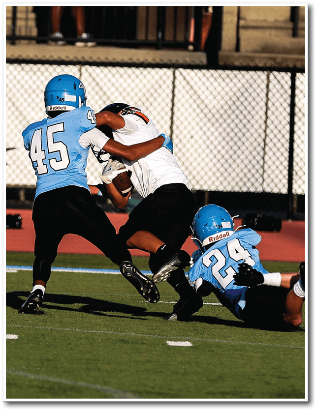 Two people in blue helmets and jerseys tackle a person in a white jersey holding a football on a grassy field. Description generated by AI