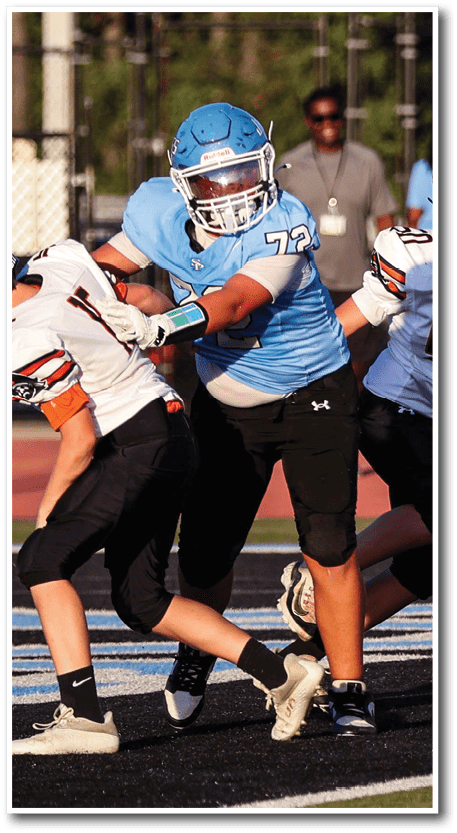 A person wearing a blue football uniform and helmet is blocking another person in a white and black football uniform during a game on an outdoor field. Description generated by AI