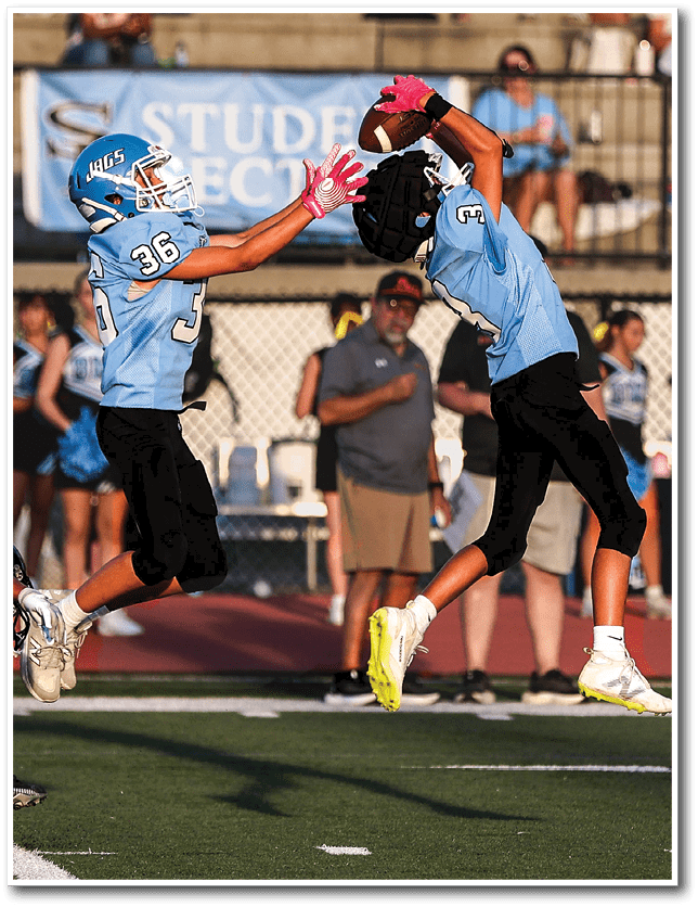 Two people in light blue football uniforms and pink gloves are actively reaching to catch a football during a game on a field with spectators in the background. Description generated by AI
