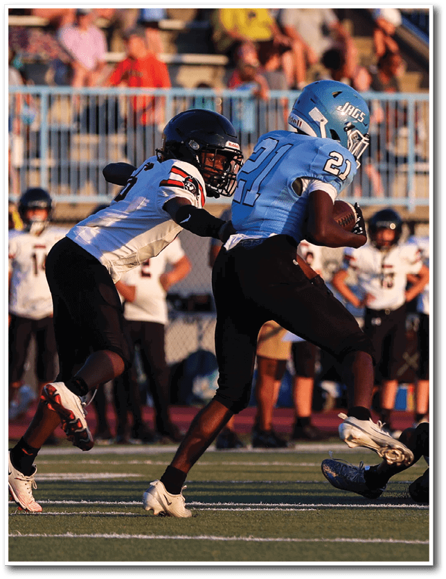 Two people in football uniforms are engaged in a play on the field with one person holding the ball and the other attempting to tackle. Description generated by AI
