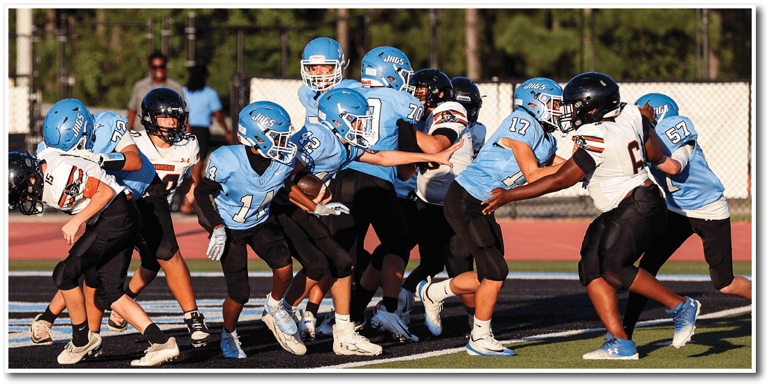Children wearing helmets and football uniforms are actively engaged in a youth football game on a field with a fence and trees in the background. Description generated by AI