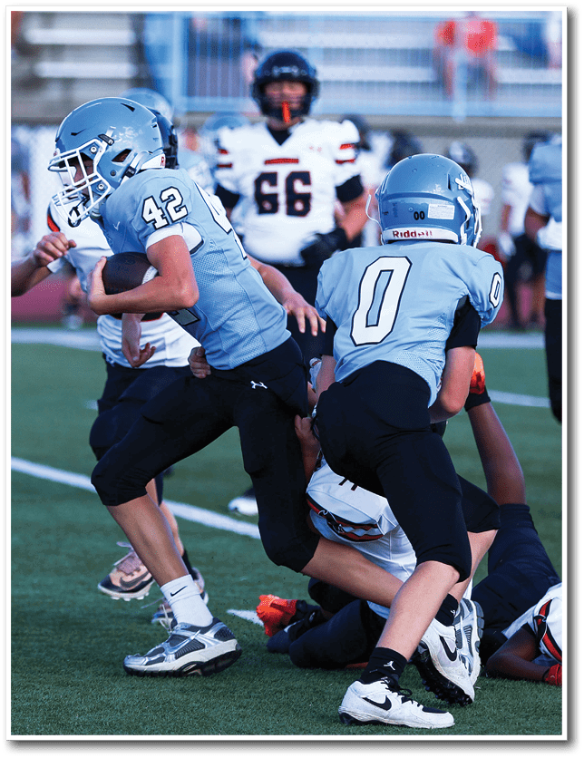 People wearing football uniforms are engaged in a game with one person holding the ball and others preparing to block or tackle. Description generated by AI