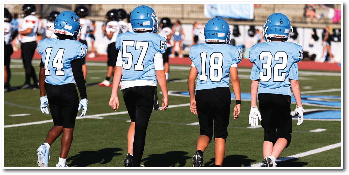 Four people wearing light blue sports jerseys with numbers on the back are walking on a grassy field. Description generated by AI