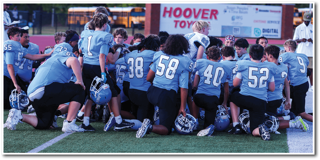 A group of people wearing light blue sports jerseys with numbers are kneeling on a field with their helmets placed on the ground in front of them. Description generated by AI