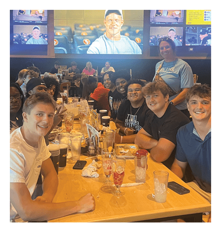A group of people seated in a dark room watch a large screen displaying a person wearing a light blue shirt and a white cap speaking. Description generated by AI