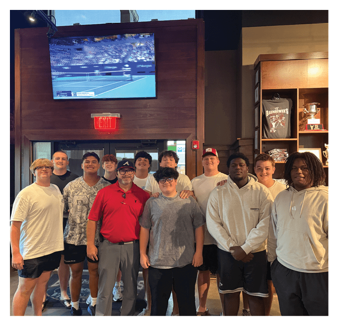 A group of ten people wearing casual clothing are standing closely together indoors near a wooden wall with a mounted television screen displaying a sports event. Description generated by AI