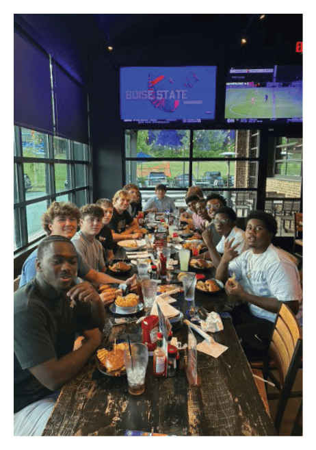 A group of people sitting around a table in a room with large windows watching two televisions mounted on the wall, one showing a soccer game and the other displaying a graphic with the text “BOISE STATE." Description generated by AI