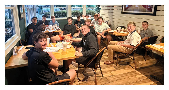 Several people wearing casual black clothing are sitting around a wooden table in a room with large windows and green walls, appearing engaged in a group discussion or meeting. Description generated by AI