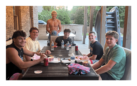 Three people are gathered outdoors under string lights, with one person shirtless holding a basketball and the other two sitting at a table with drinks and snacks. Description generated by AI