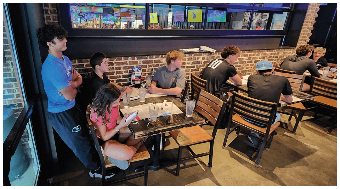 Several people are seated and standing around tables in a casual indoor setting with multiple TV screens mounted on the wall. Description generated by AI