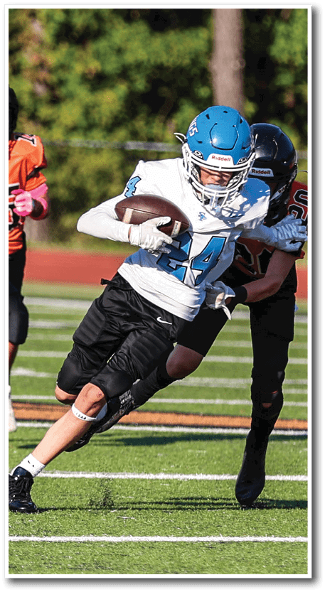 A person wearing a blue helmet and white jersey is running with a football while being pursued by two people in black and orange jerseys on a grassy sports field. Description generated by AI