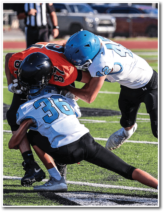 Three people wearing football helmets and uniforms are engaged in a tackle on a grassy field during a game. Description generated by AI