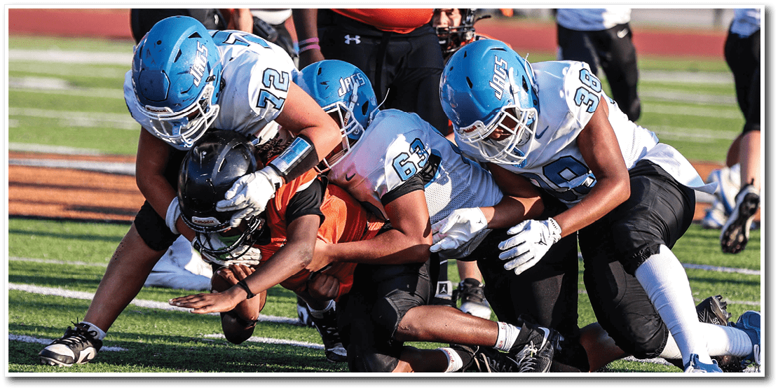 People wearing football uniforms are gathered on a field with some standing and others crouching, engaged in a game or practice. Description generated by AI