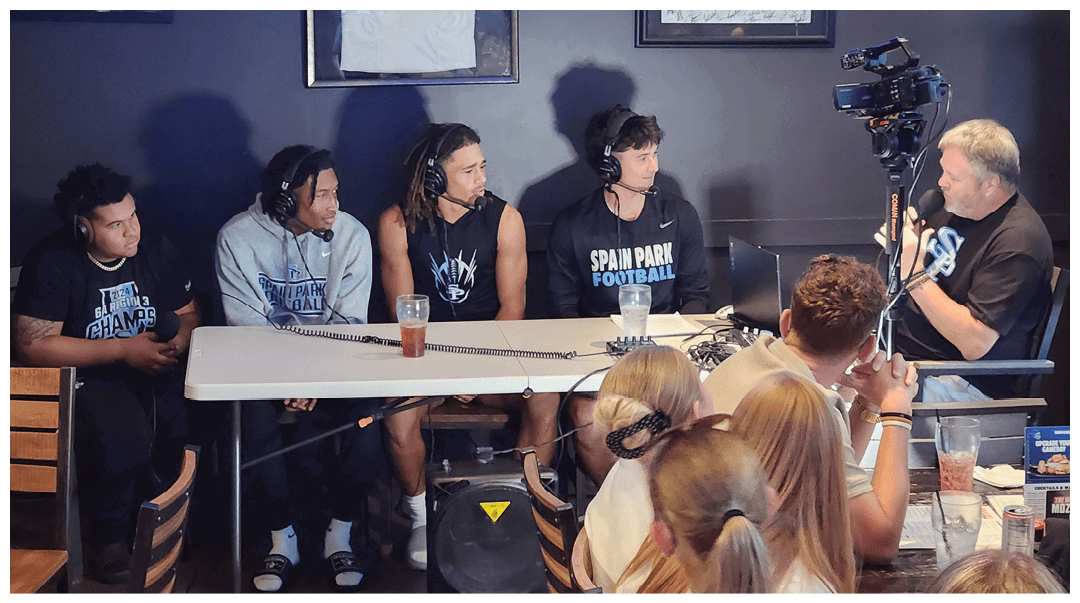 Four people wearing headphones and casual sportswear sit at a table with microphones and drinks while another person speaks into a microphone in front of a camera in a dimly lit room with an audience watching. Description generated by AI