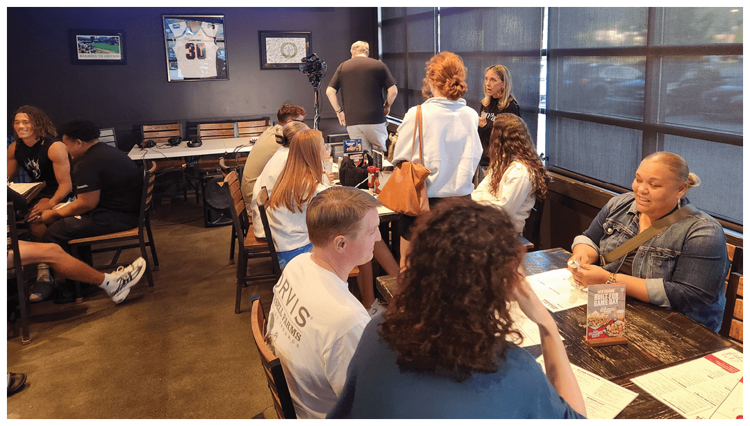 People are sitting and standing in a casual dining area with large windows and multiple TV screens showing a sports game. Description generated by AI