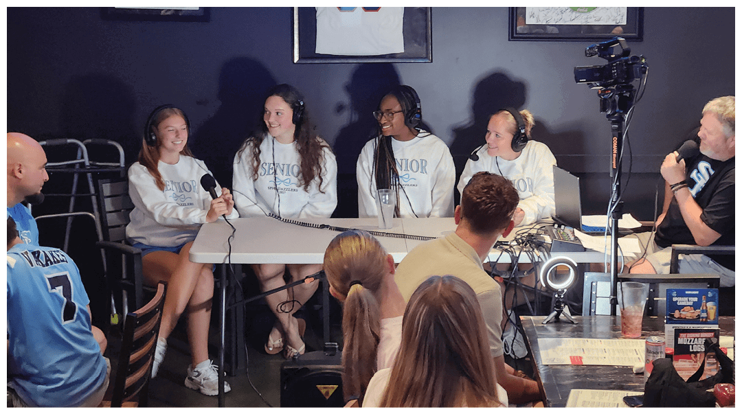 Four people wearing matching “Senior" sweatshirts sit at a table with microphones while two people operate a camera and audio equipment in a dark room with framed sports jerseys on the wall. Description generated by AI