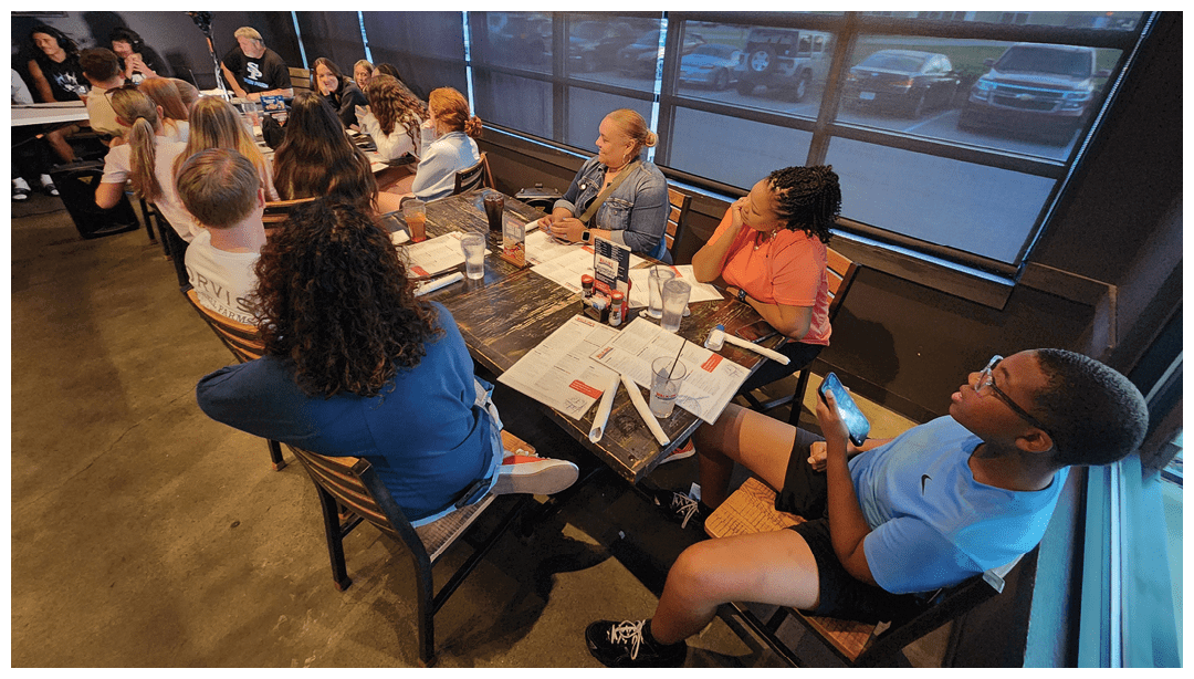 A group of people sitting around a table in a restaurant, engaged in conversation with menus, drinks, and condiments on the table. Description generated by AI