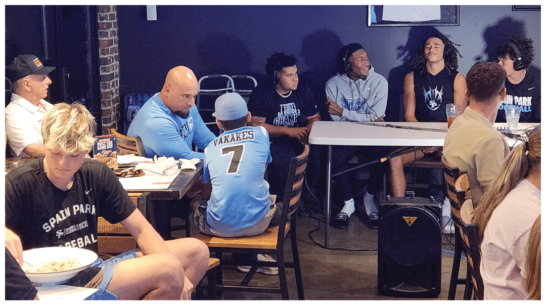A group of people wearing casual clothing and sports jerseys are seated around tables in a room, engaged in conversation and eating. Description generated by AI