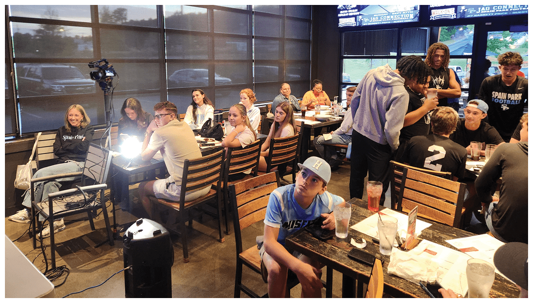 A group of people sitting and standing around tables in a casual indoor setting with large windows and some recording equipment nearby. Description generated by AI